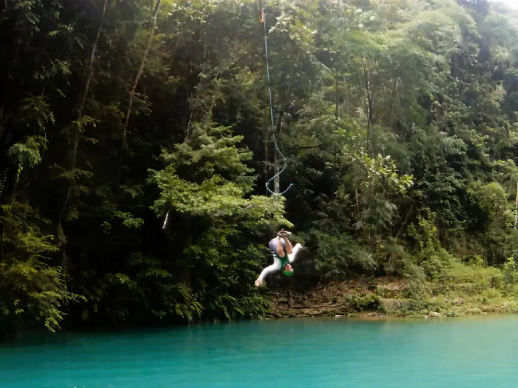 Person swinging on a rope over turquoise water surrounded by lush trees.