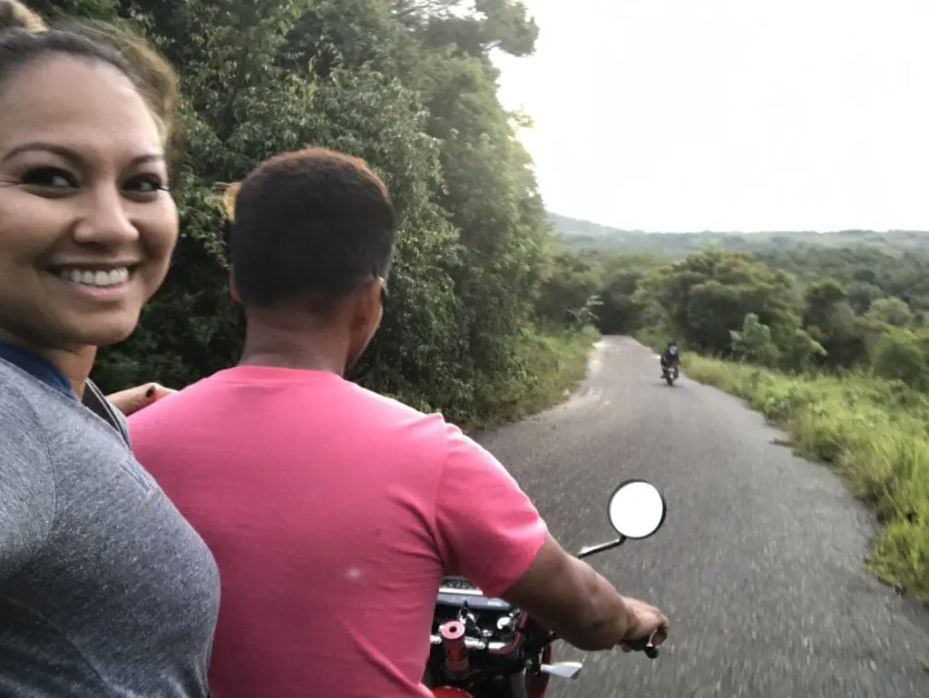 Two people riding a motorcycle on a winding road surrounded by greenery.