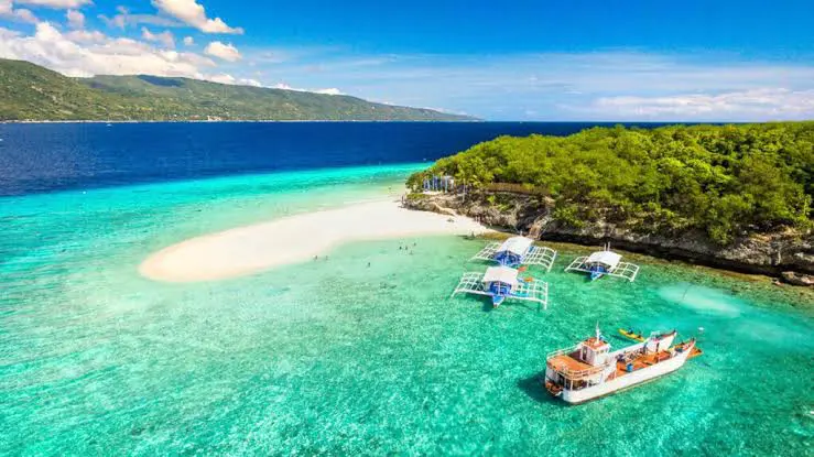 Crystal-clear turquoise waters and sandy beach with boats near lush greenery.