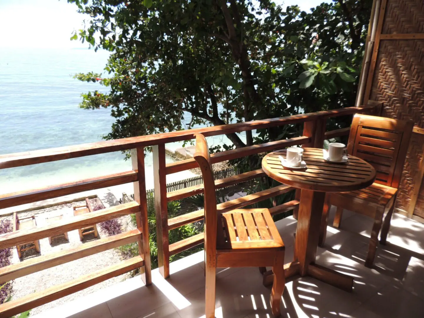 Wooden balcony with chairs overlooking the sea under leafy shade.
